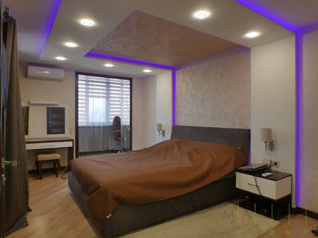 на фото мебель для спальни, кровать оббитая мягкой тканью и прикроватная тумбочка от стены и по потолку идет подсветка фиолетового цвета сделанная на заказ