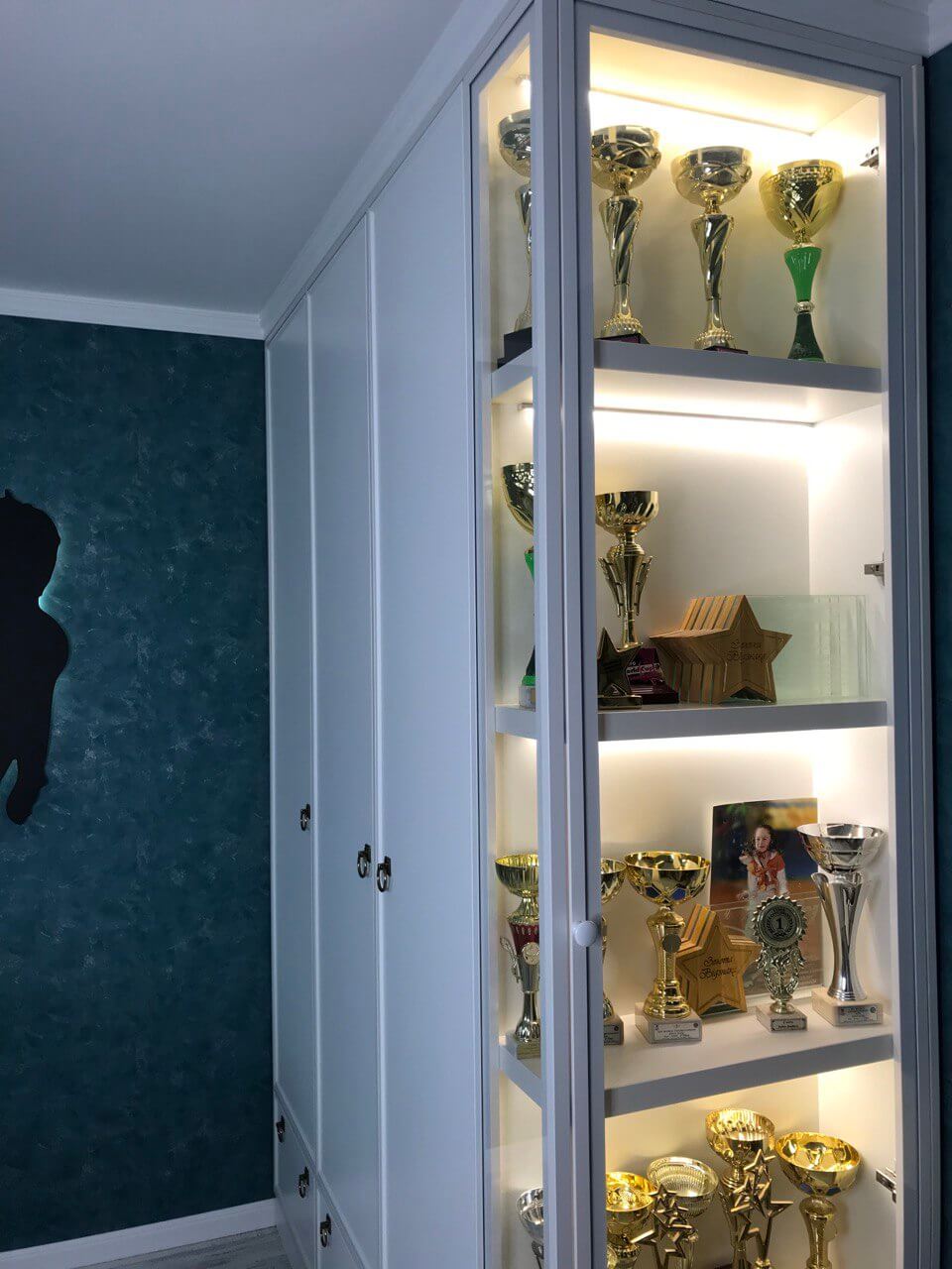 на фото шкаф в детскую комнату, справа сделан ряд сверху донизу с подсветкой для спортивных наград. Правая часть шкафа с наградами на первом плане